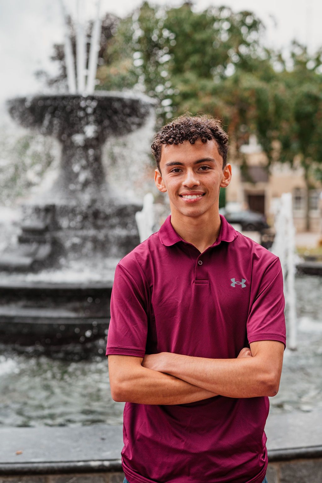 Young man in maroon Under Armour polo smiles with arms crossed in front of a decorative fountain outdoors.