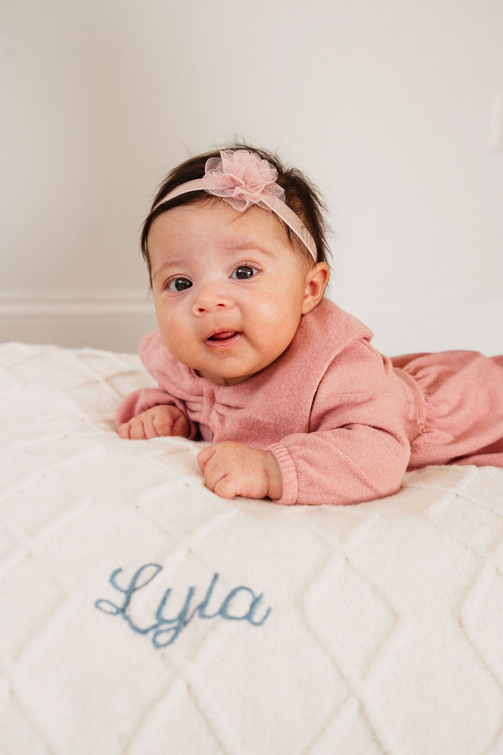 Baby girl named Lyla in pink outfit and floral headband doing tummy time on white quilted blanket.