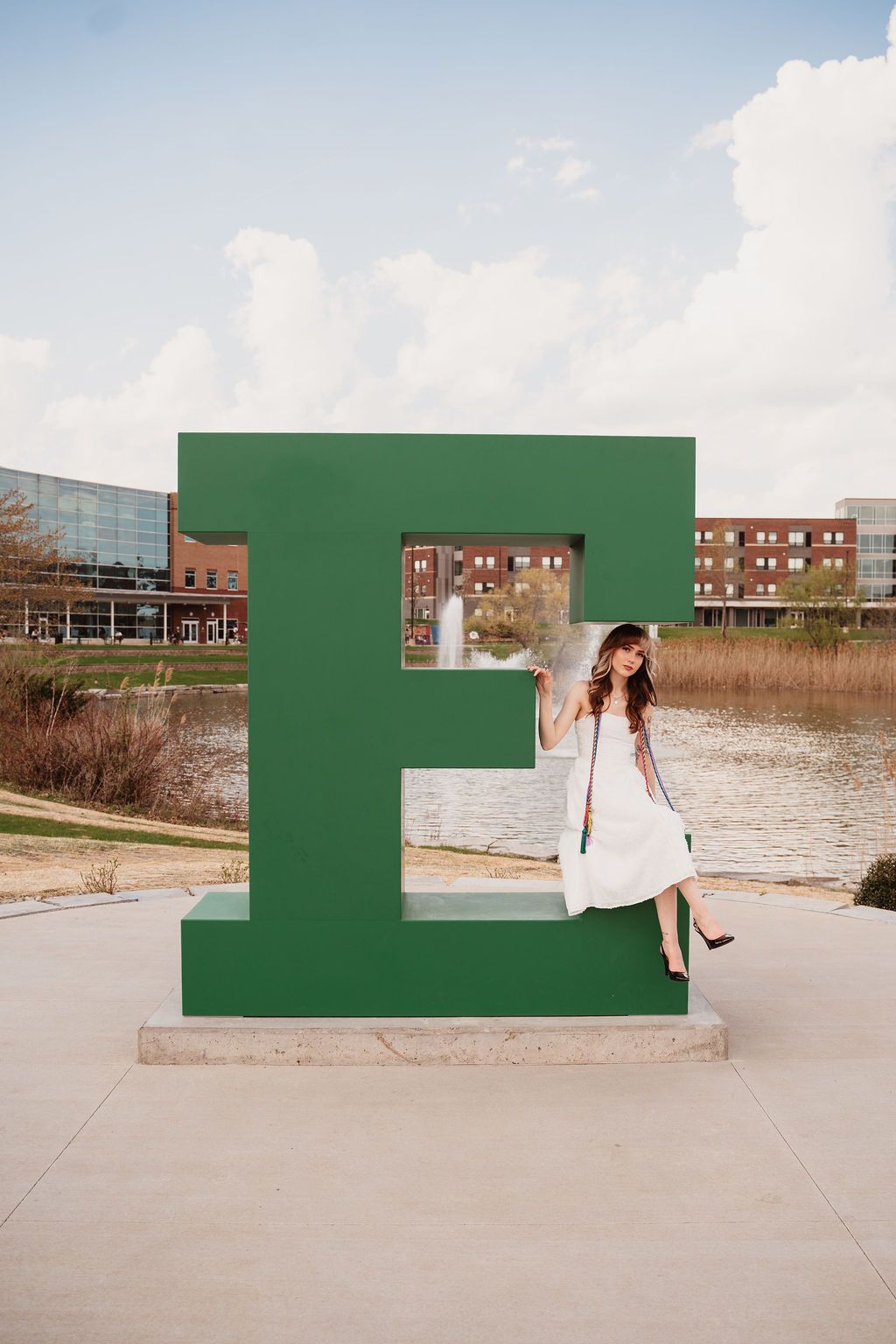 Woman in white dress posing beside large green letter E sculpture near a pond on a college campus.