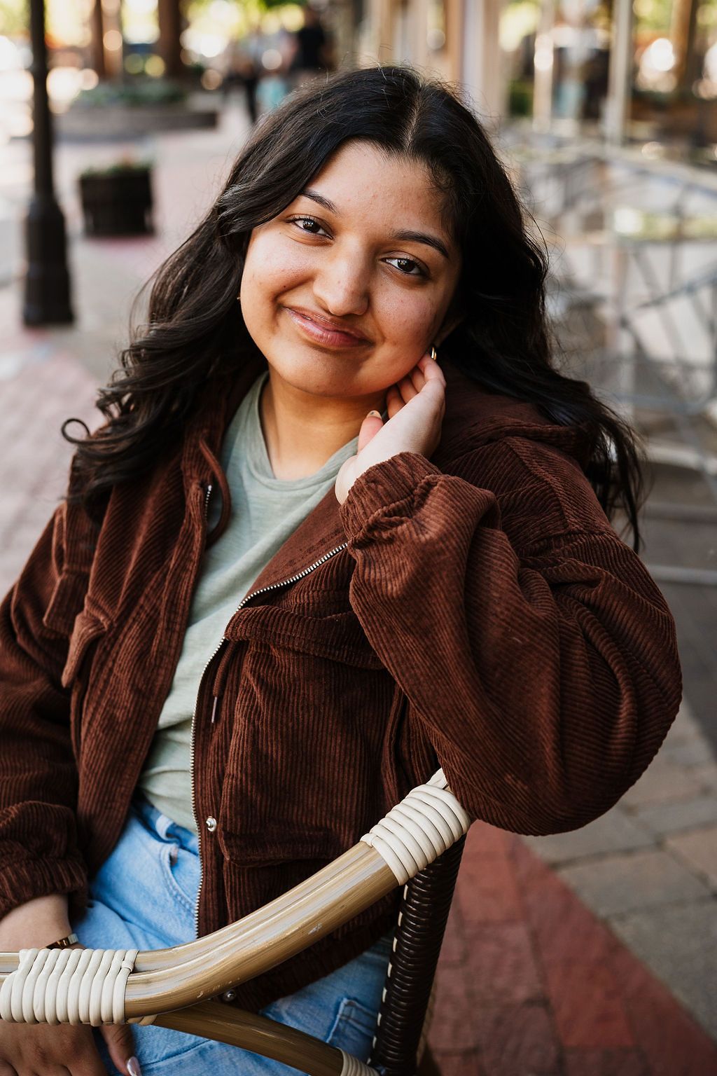 Young woman with dark curly hair smiling, wearing a brown corduroy jacket, seated outdoors on a wicker chair.
