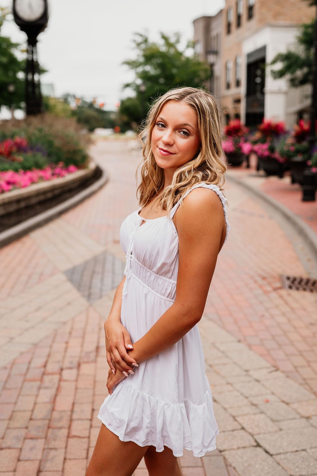 Young woman in white sundress posing on brick pedestrian walkway with colorful flower beds and buildings.