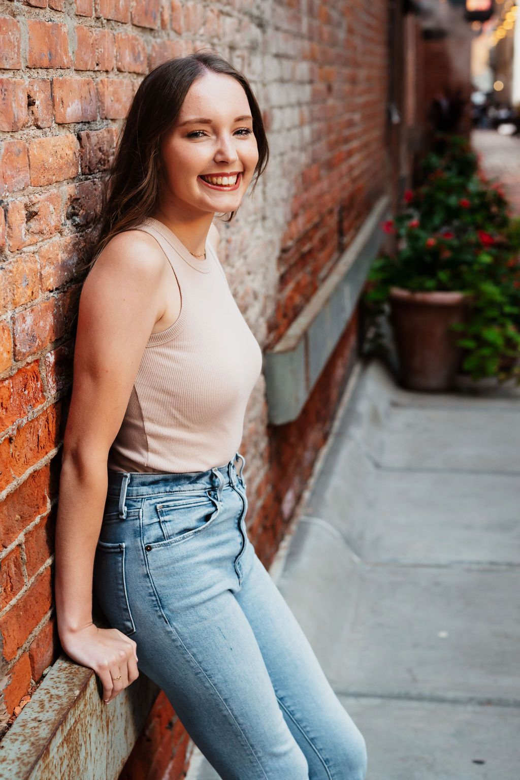 Young woman smiling in jeans and beige tank top, leaning against a red brick wall on a charming urban sidewalk.