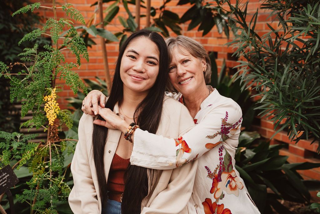 Two women smiling and embracing outdoors among lush green plants near a brick wall.