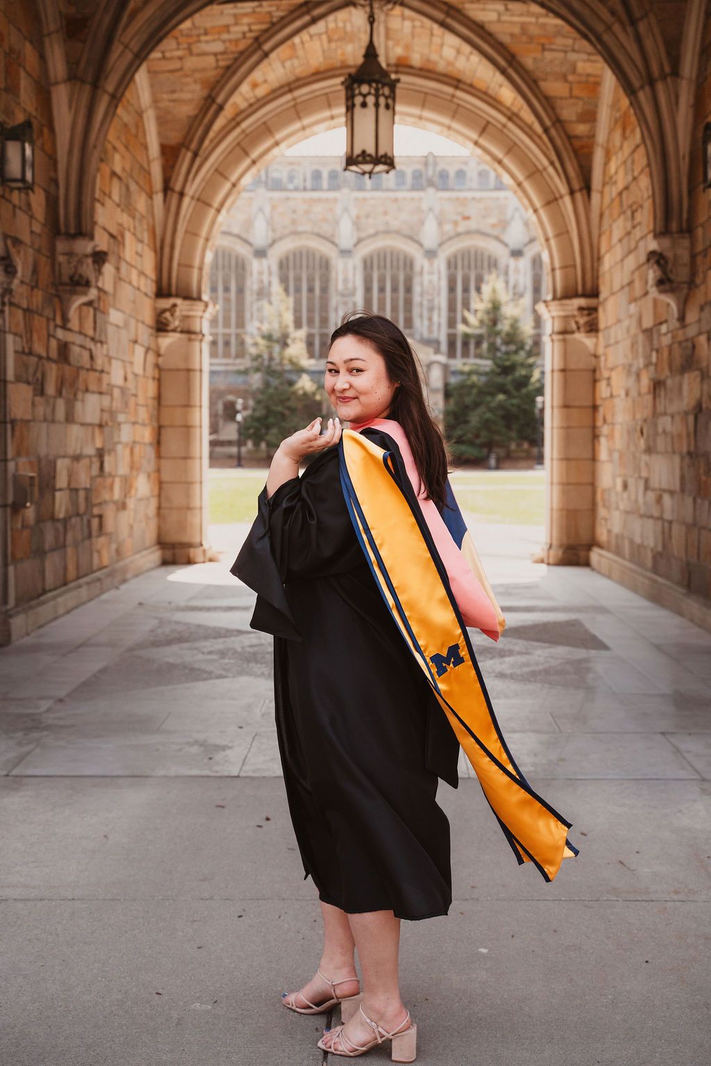 Graduate in black gown with gold Michigan stole poses under stone archway on U of M campus.