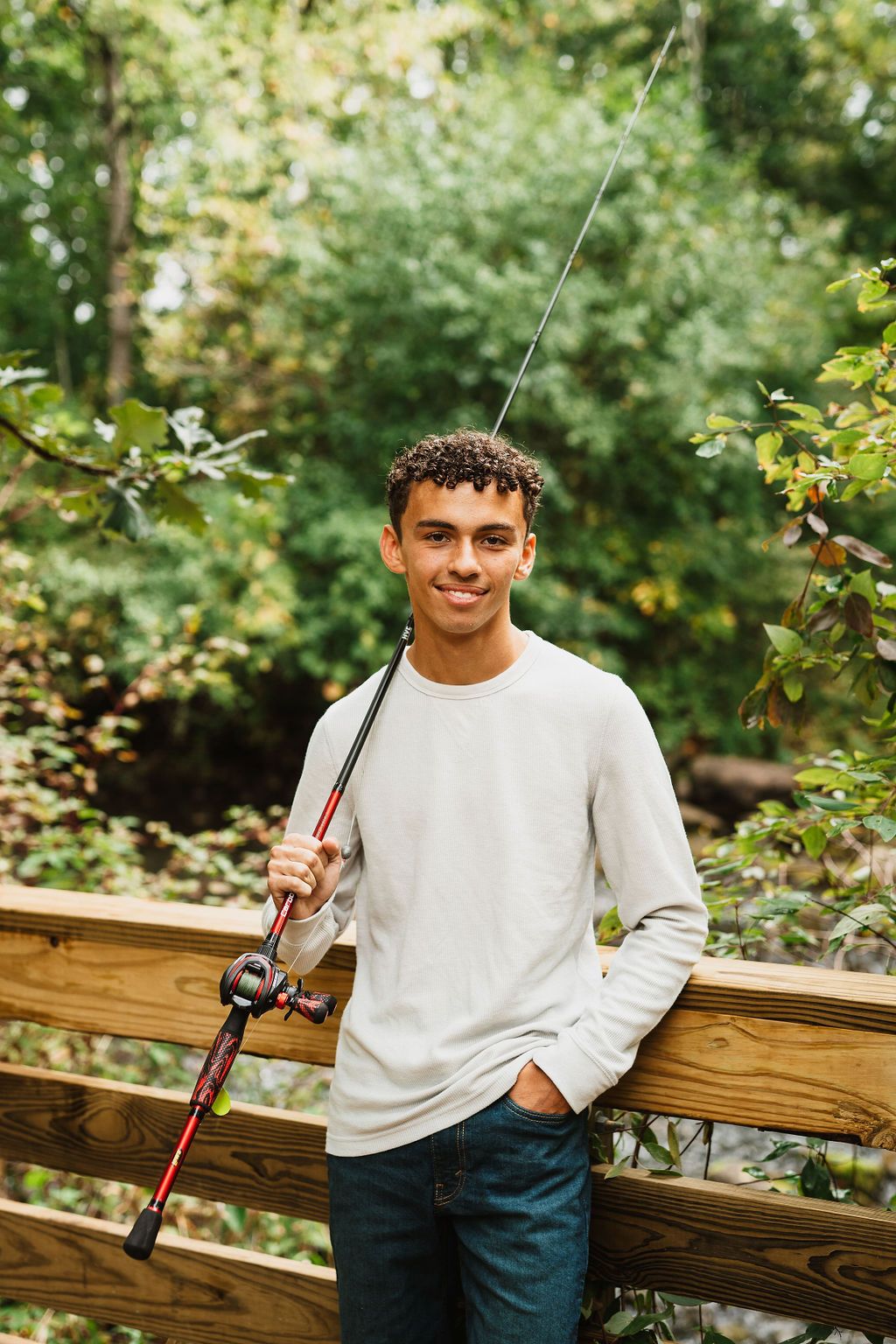 Young person in white sweater and jeans sitting on wooden railing in outdoor nature setting.