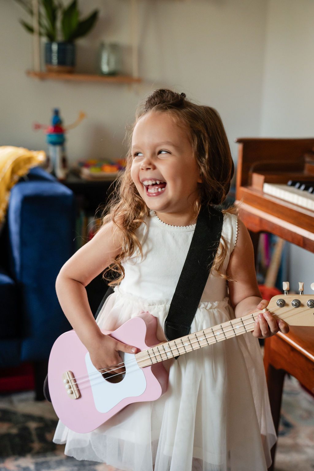 Joyful little girl in white dress playing pink ukulele, laughing with piano in background.