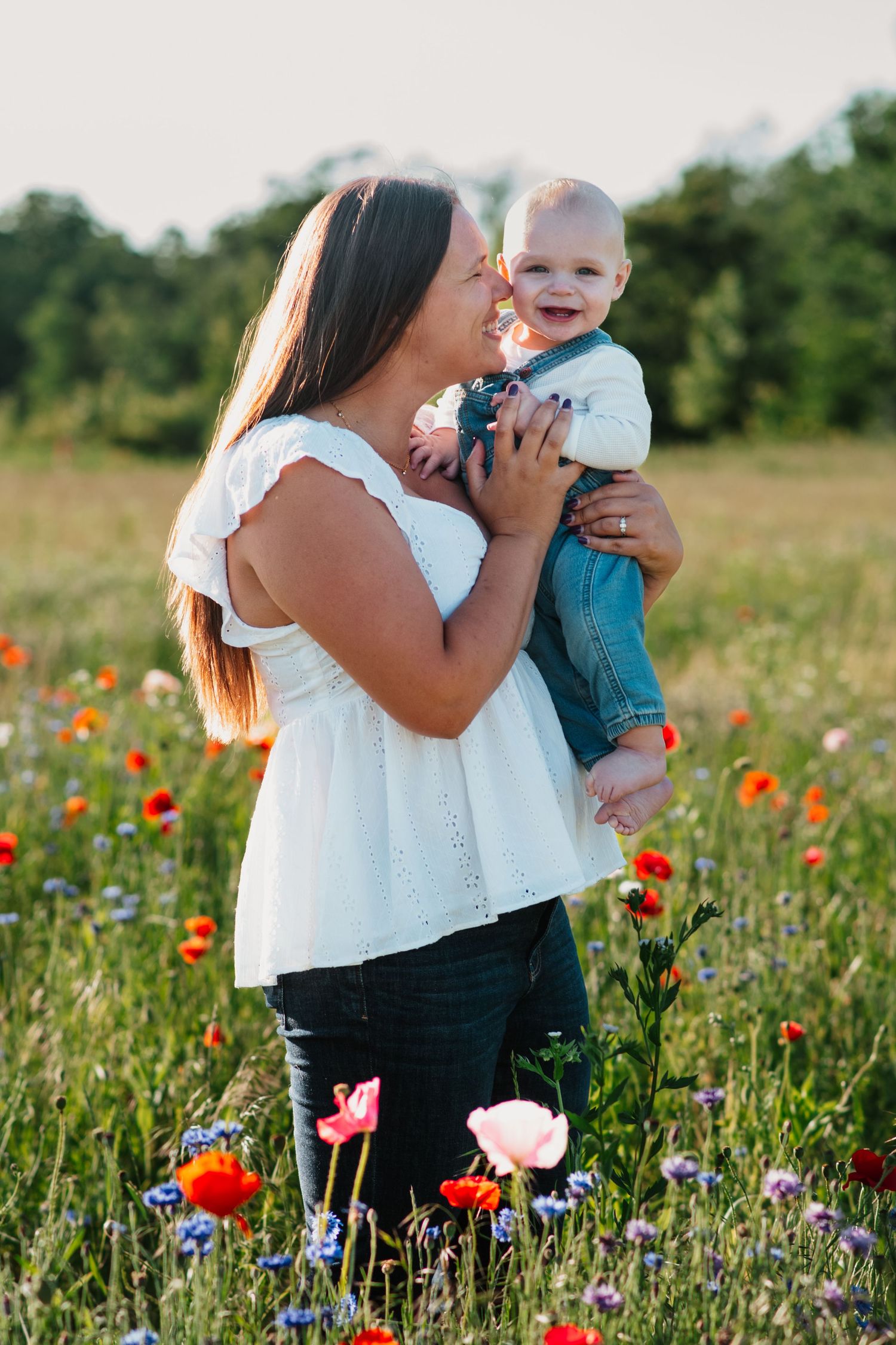Mother holding smiling baby in a vibrant wildflower field with red poppies and cornflowers on a sunny day.