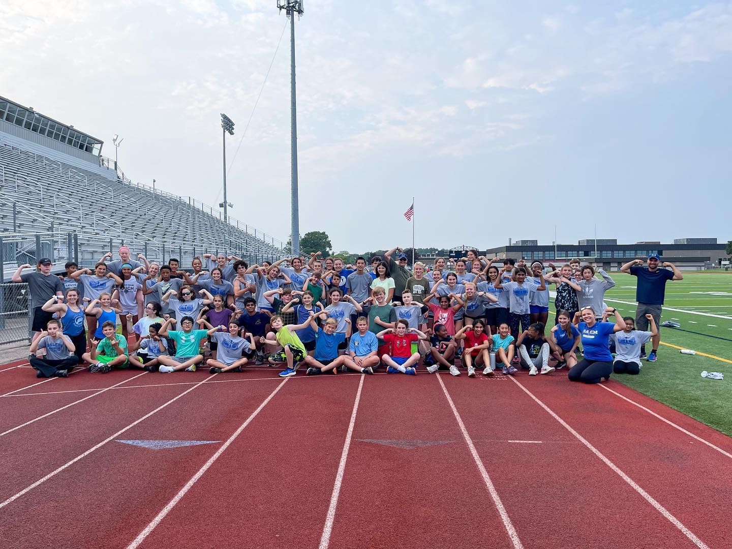 Large group of kids and coaches posing on a red track at an outdoor stadium on a sunny day.