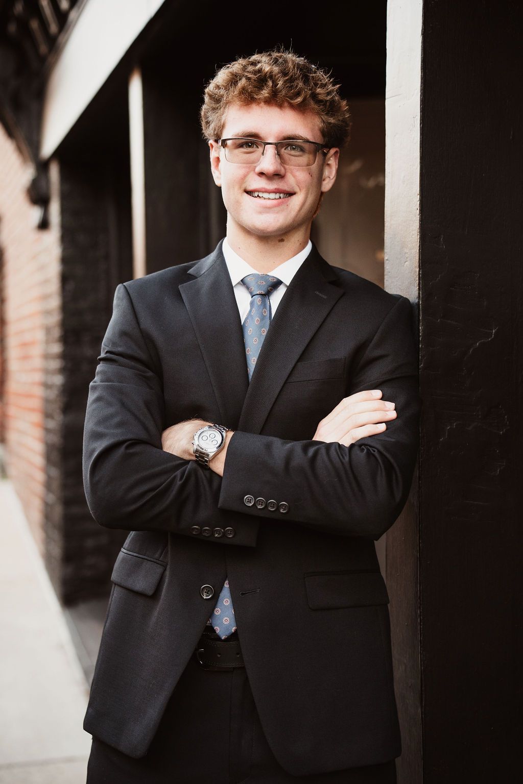 Young professional man in black suit, blue tie, and glasses smiling with arms crossed outdoors.