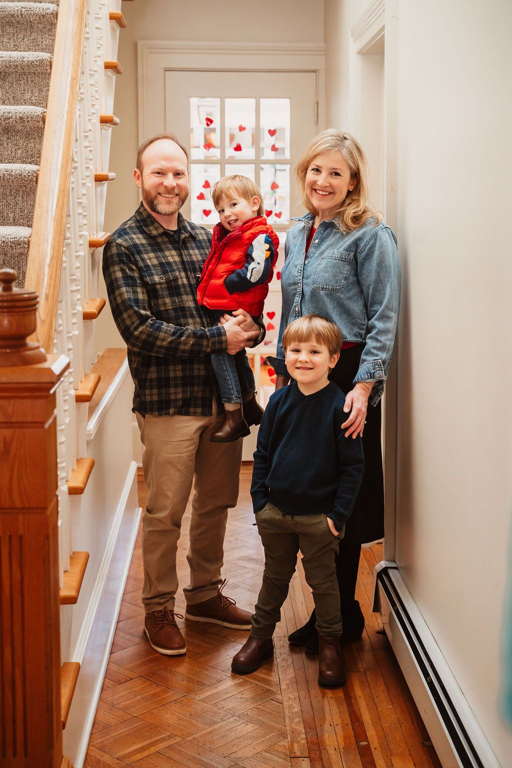 A happy family of four poses in a cozy home entryway decorated with Valentine's Day hearts, featuring two young boys.