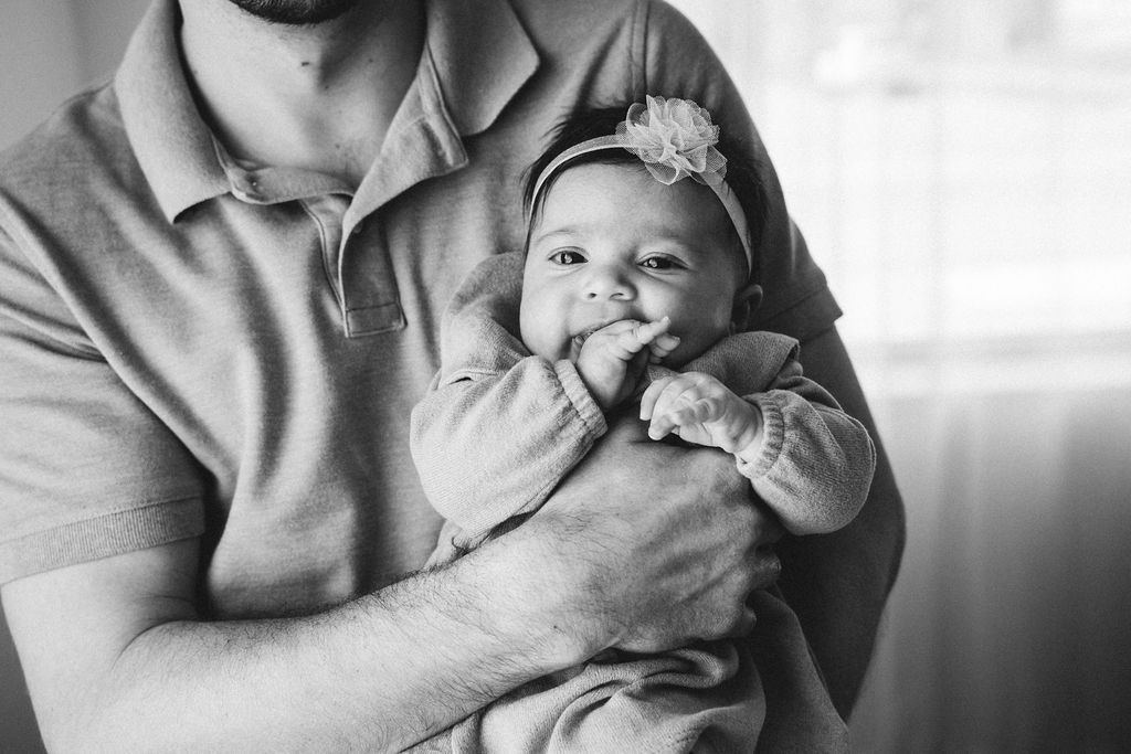 Black and white photo of a father tenderly cradling his newborn baby girl wearing a floral headband.