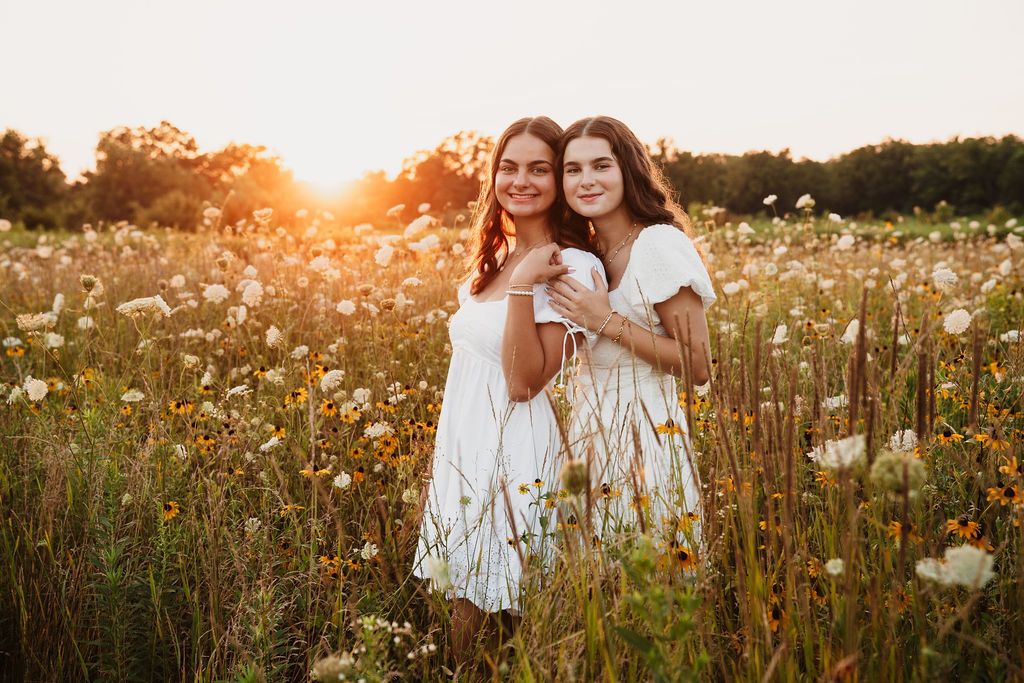 Two women posing in a wildflower field at golden hour with warm sunlight creating a dreamy atmosphere.
