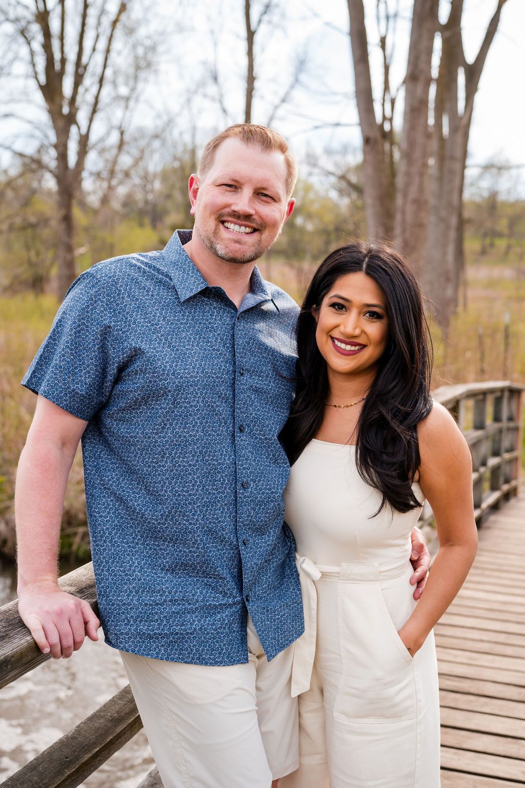 A happy couple in coordinated blue and white outfits pose together on a wooden bridge during an outdoor photoshoot.