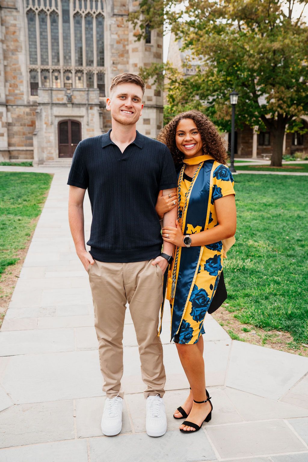 Two students stand together on a university campus pathway with Gothic architecture and trees in the background.