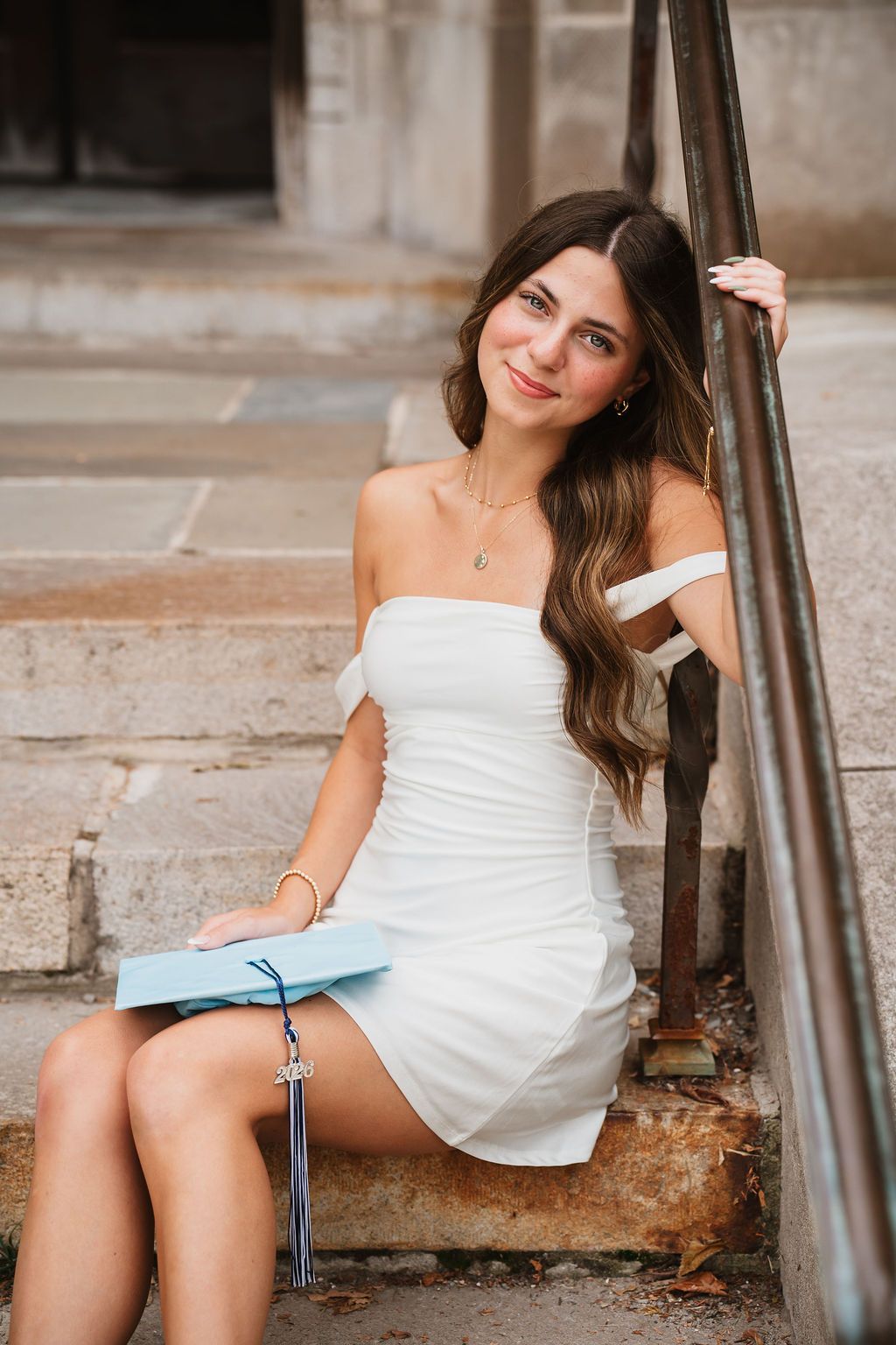 A person in a white off-shoulder dress sits on stone steps with a graduation cap nearby.