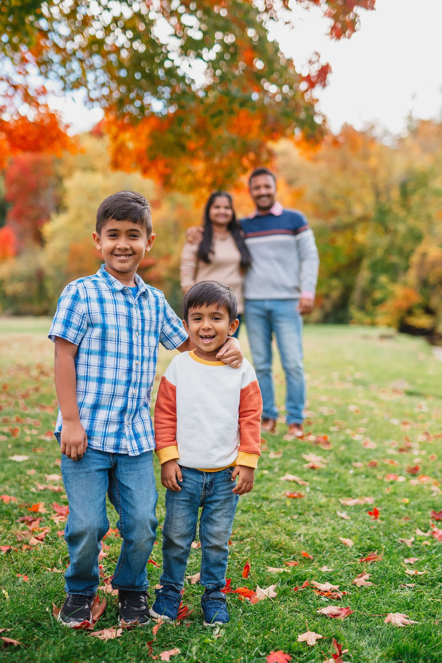 Family enjoying a fall day outdoors with autumn foliage and fallen leaves on green grass. Northville Hines park fall mini sessions