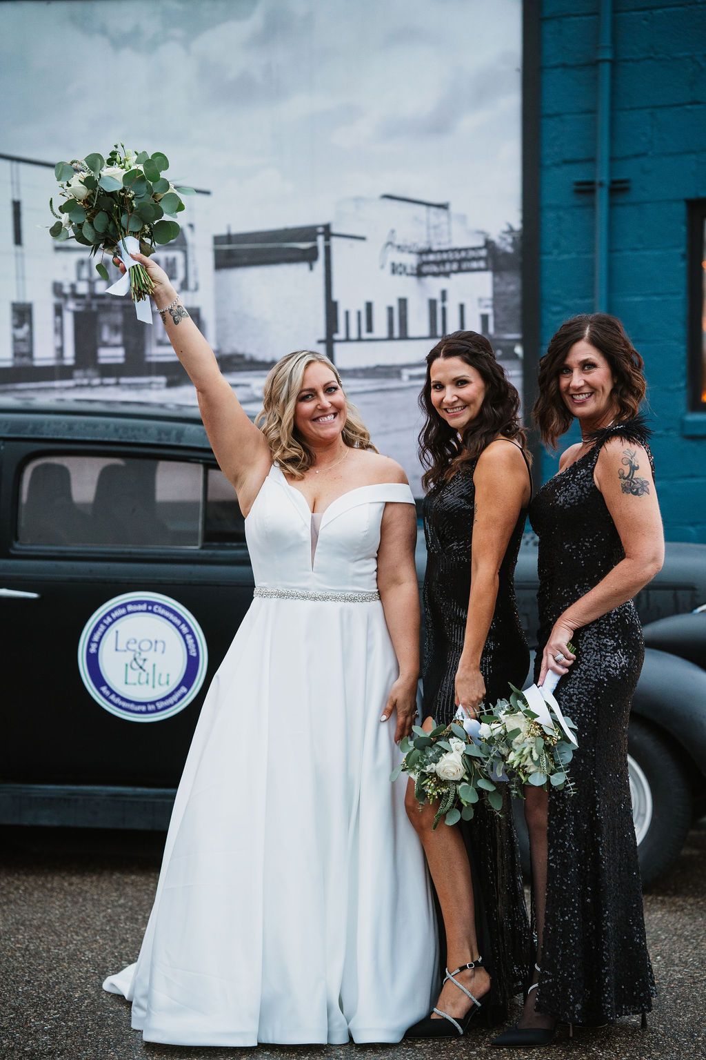 Wedding party in white dress and black sequin gowns pose against blue wall.