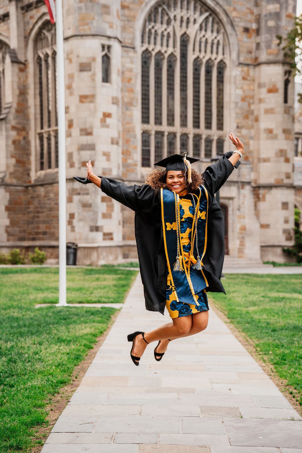 A graduate in academic regalia jumps joyfully outside a gothic church building during a celebratory photoshoot.