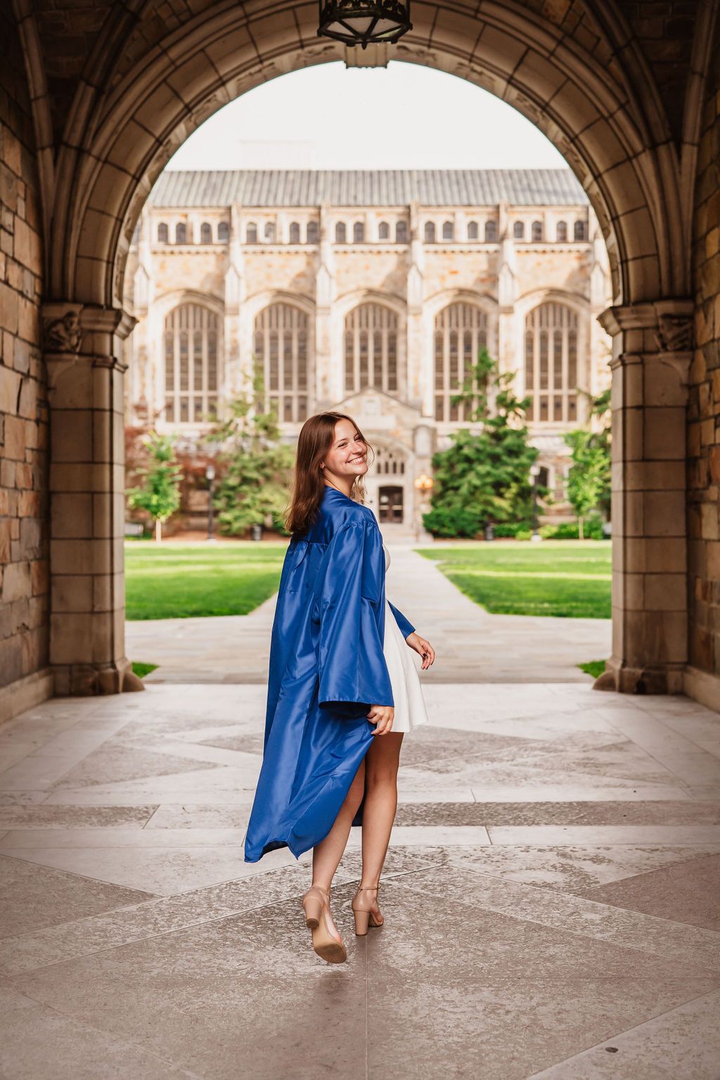 Graduate in blue cap and gown twirls under historic stone archway with collegiate gothic building in background.