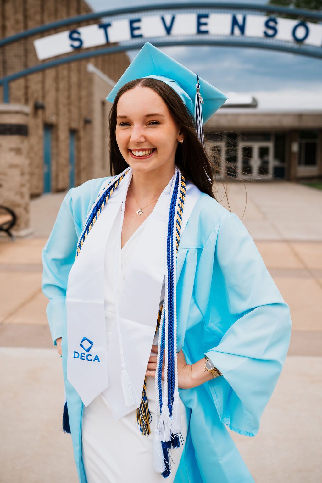 Graduate in light blue cap and gown smiles while posing for graduation photos outside a brick building.