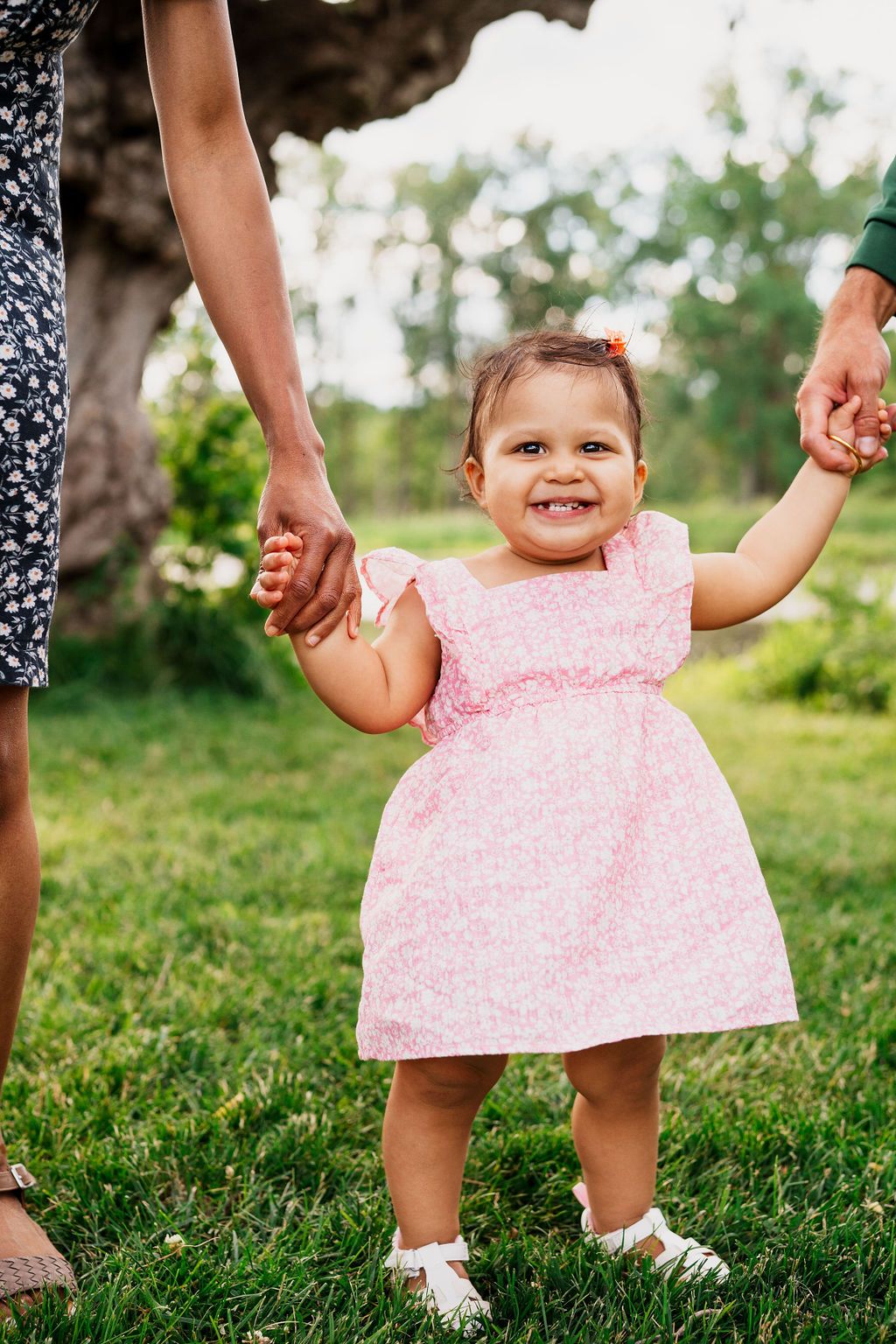 Small child in pink polka dot dress holds hands with adults while standing on grass in summer sunlight.