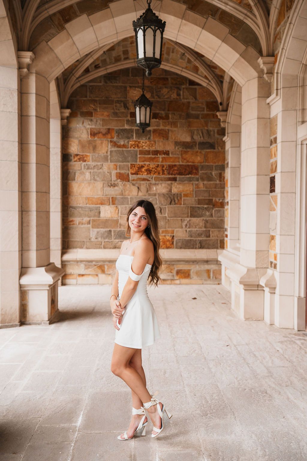 A woman in a white dress poses elegantly in a stone archway corridor with hanging lanterns.
