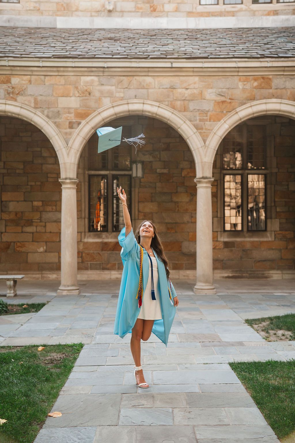 Person in light blue blazer and white dress twirls joyfully under stone archways in a historic courtyard setting.