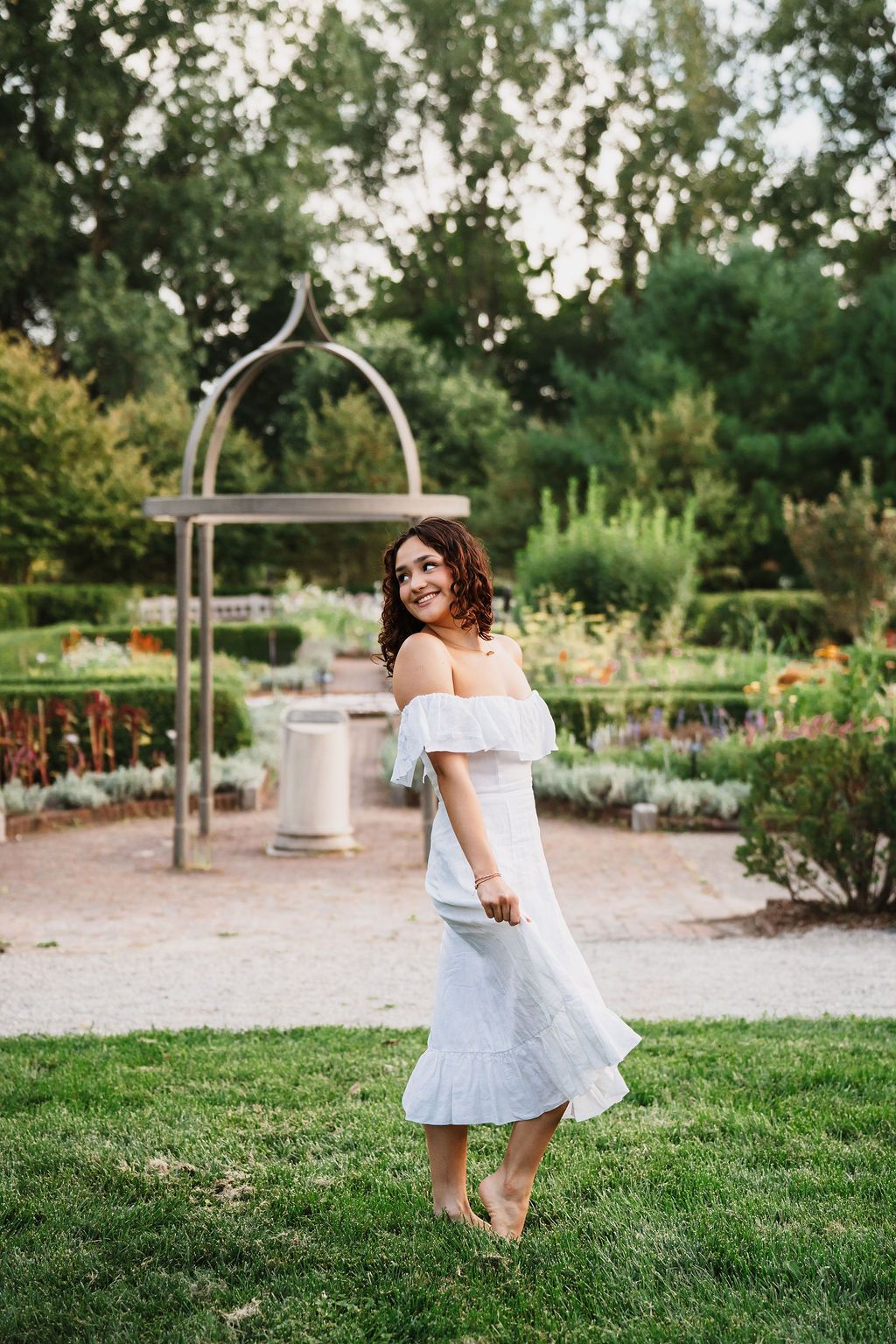 A woman in a flowing white off-shoulder dress twirls in a garden setting with an archway and greenery in the background.
