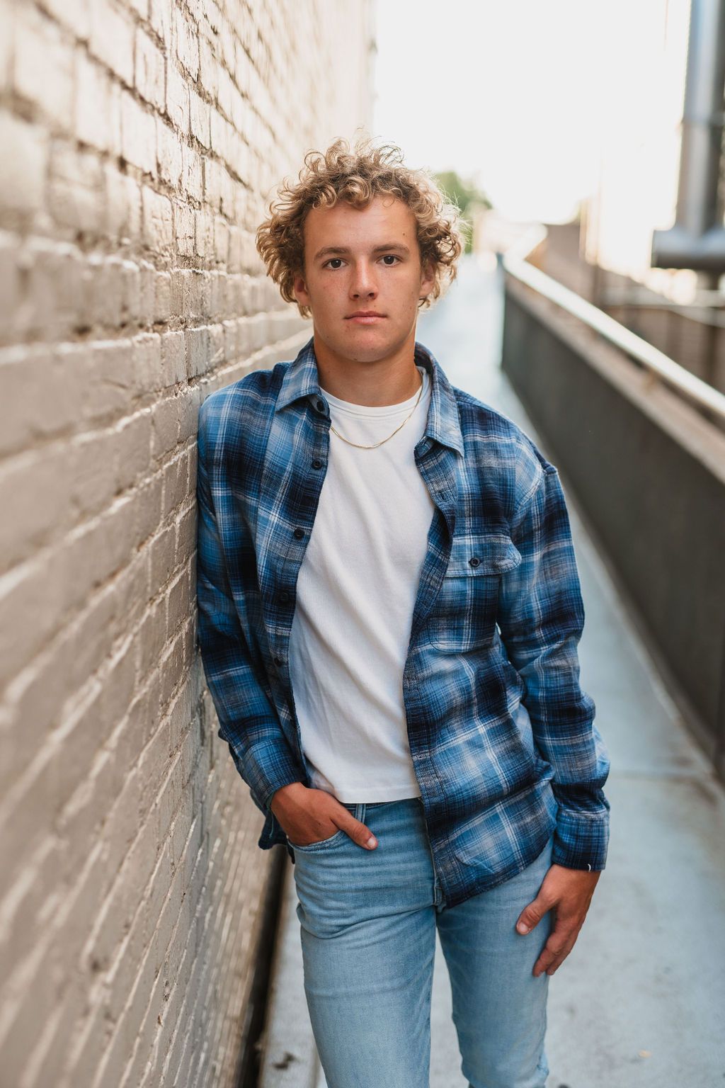 Young man with curly hair leans against brick wall wearing blue plaid flannel shirt and jeans in urban alley.