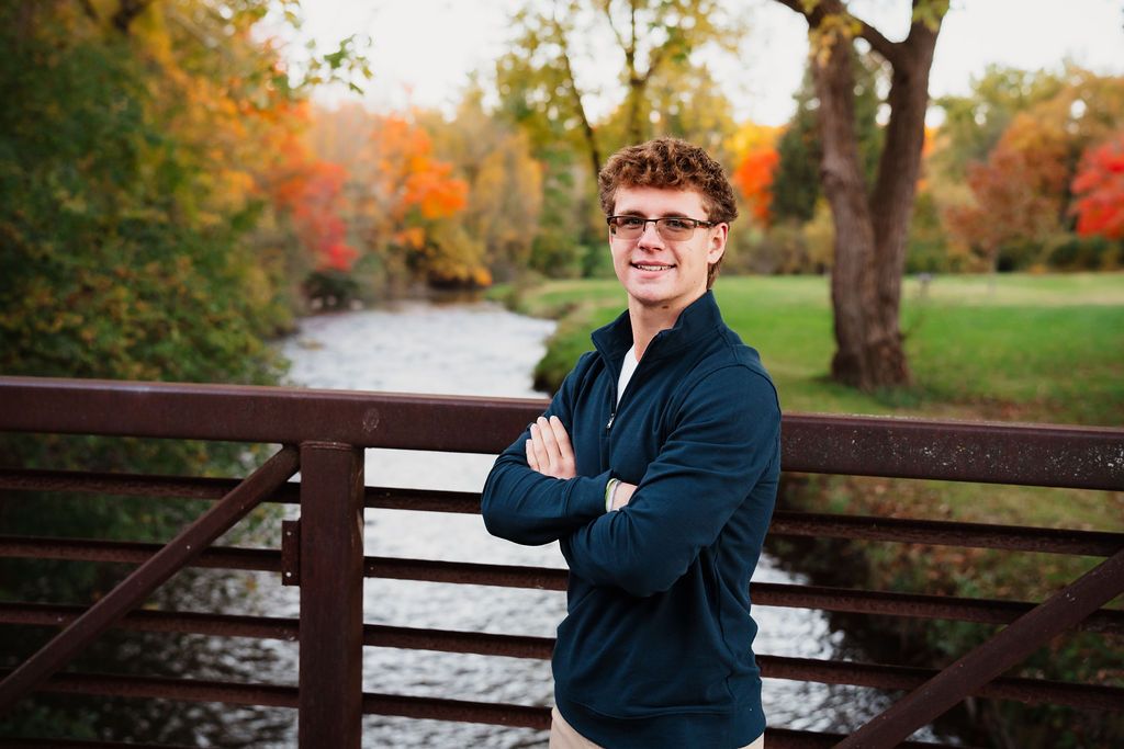 Person standing on a bridge with arms crossed, surrounded by colorful autumn foliage and a stream.