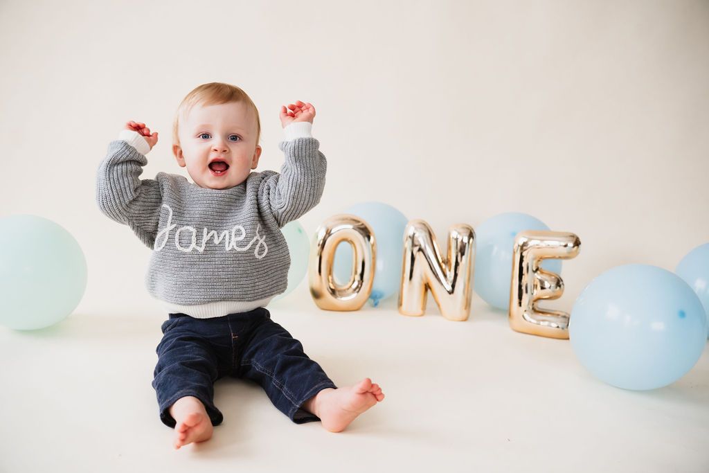 Baby boy in gray sweater sits with arms raised beside gold ONE balloons and blue balloons for his first birthday.