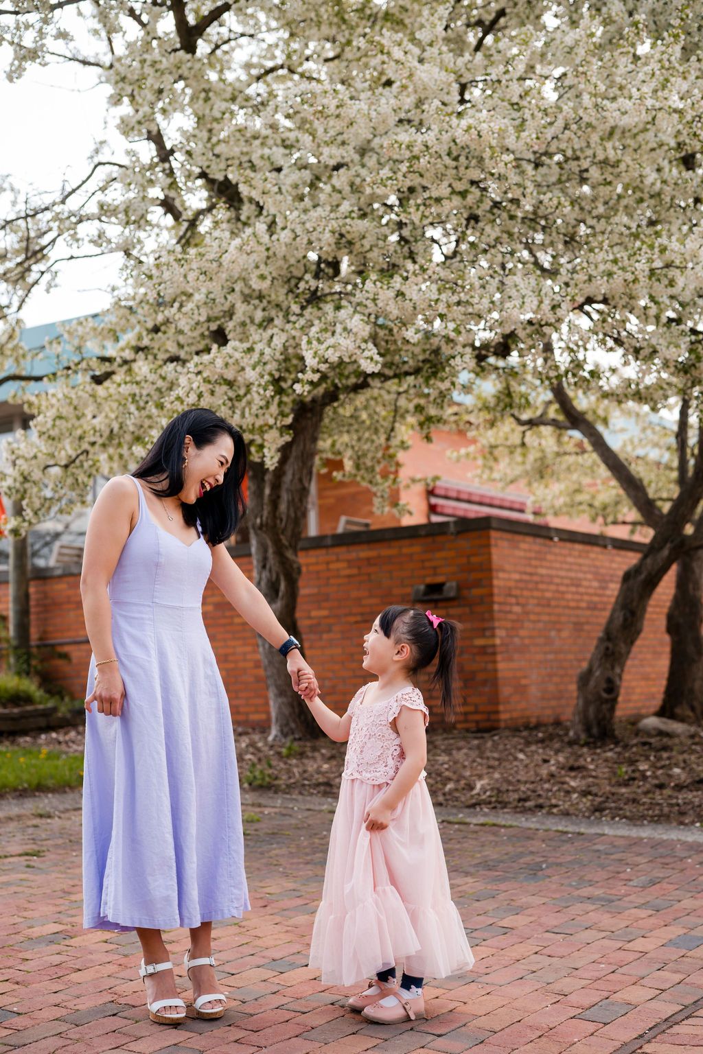 Two people in matching white and pink dresses share a tender moment under blooming cherry blossom trees on brick pavement.