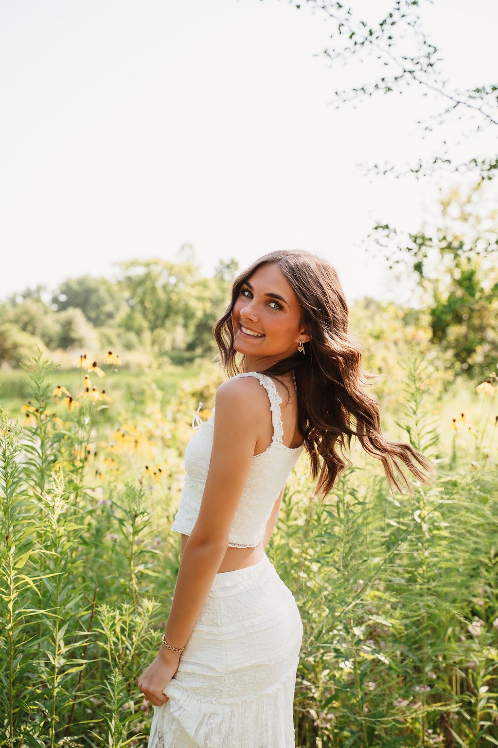 Young woman in white crop top and skirt smiling in a sunny wildflower field with lush green trees behind her.
