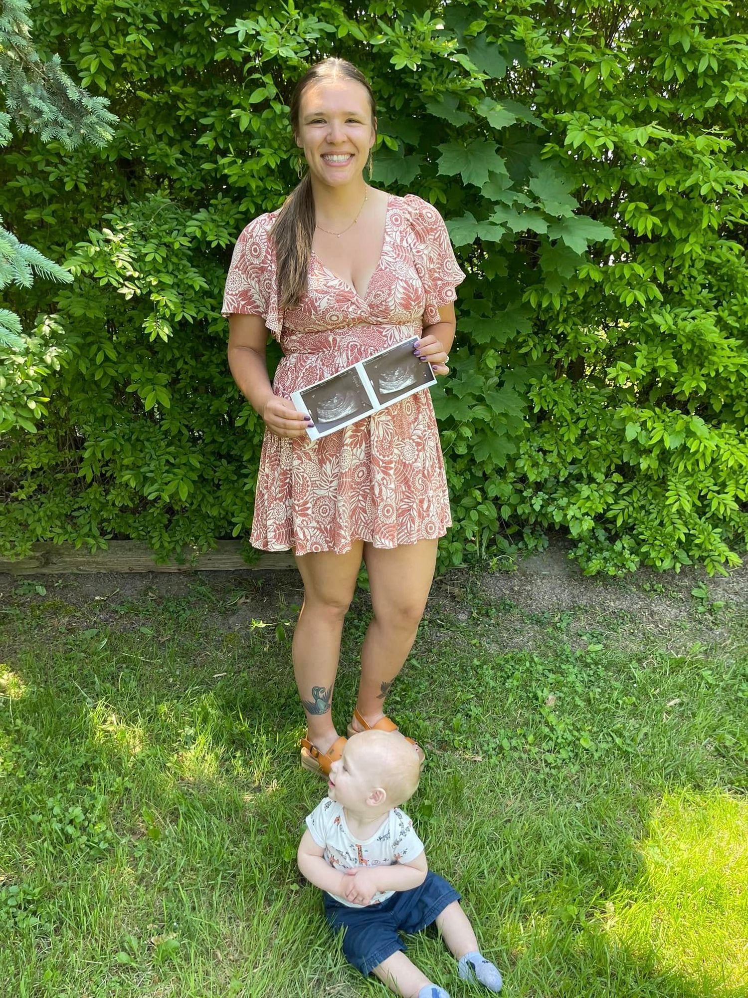 Smiling woman in floral dress holding ultrasound photos with baby sitting on grass in backyard.
