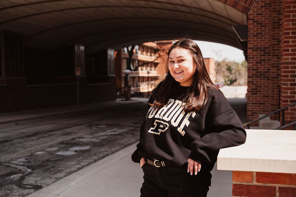 Smiling young woman wearing Purdue University sweatshirt standing under brick archway outdoors.