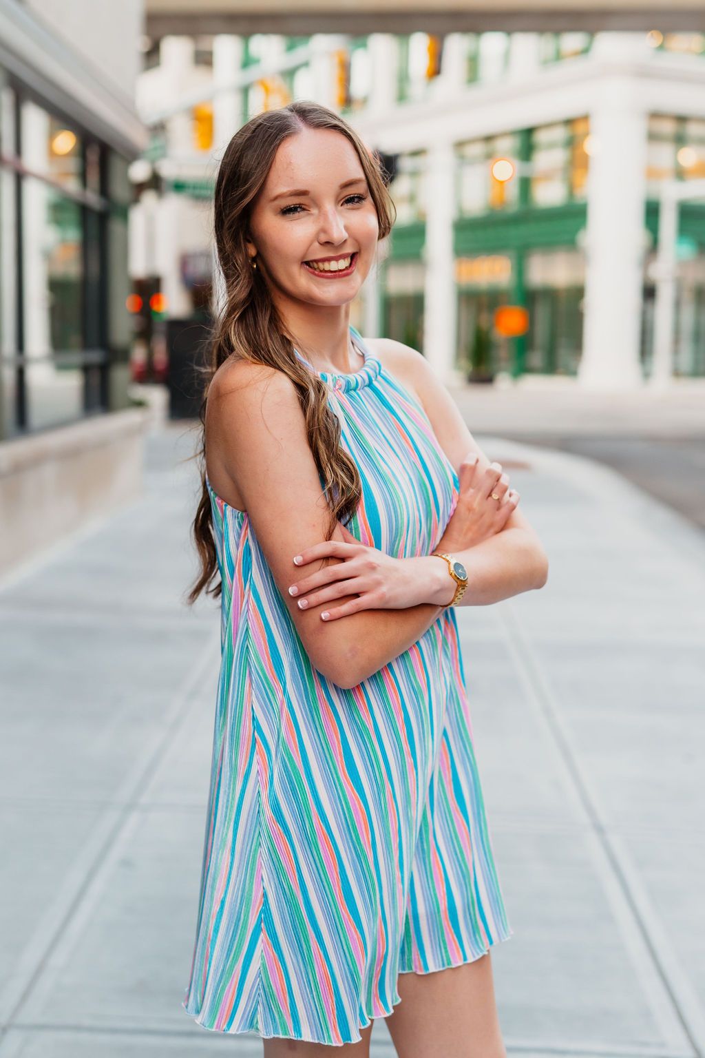 Smiling young woman in a colorful striped sundress posing with arms crossed on an urban sidewalk.