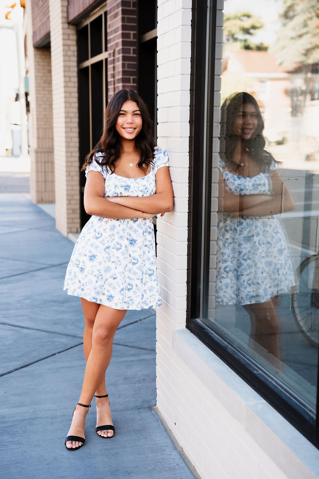 Young woman in blue floral dress leans against brick wall with her reflection in window.