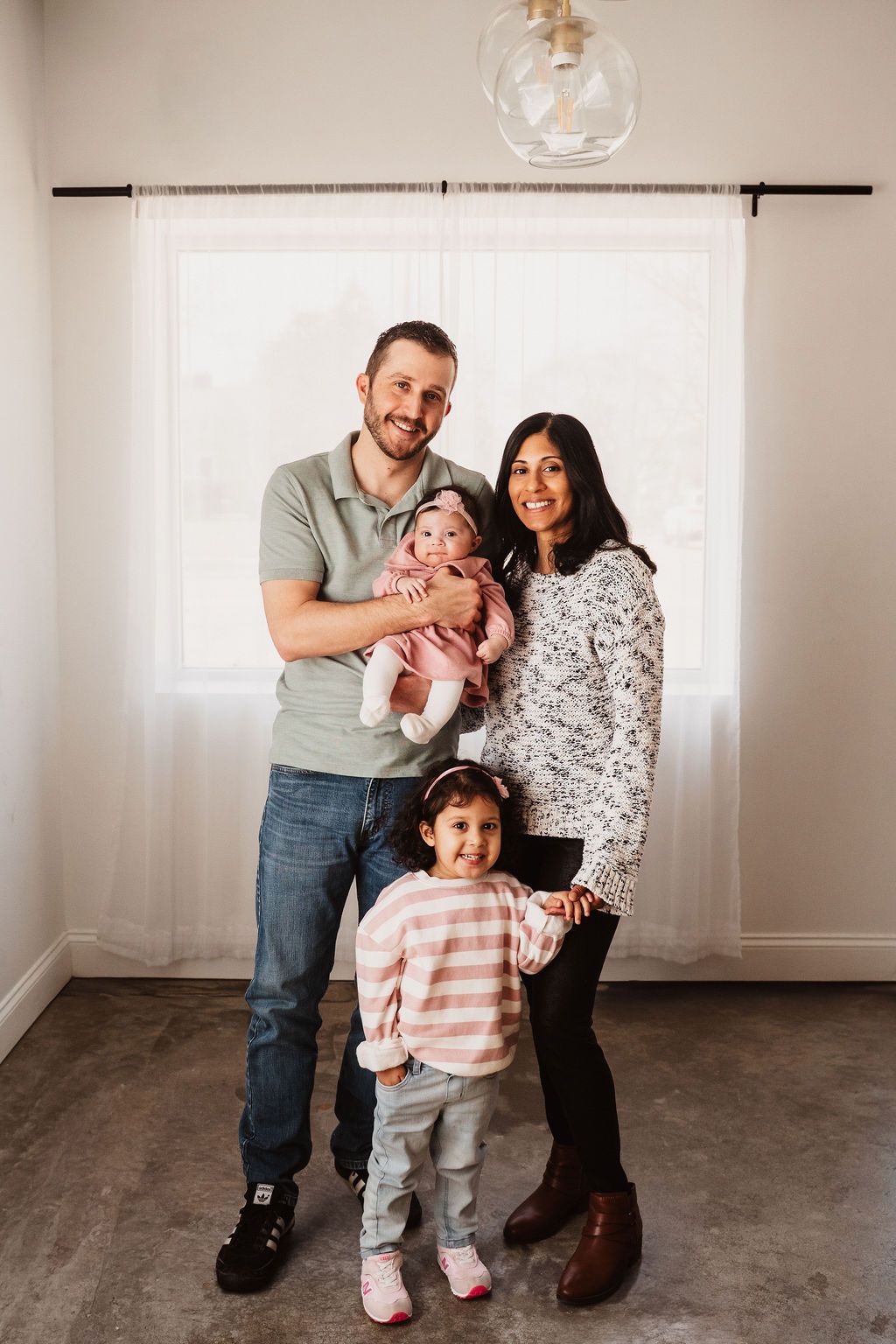 Happy family of four posing indoors, parents holding baby and young daughter smiling in bright modern room.