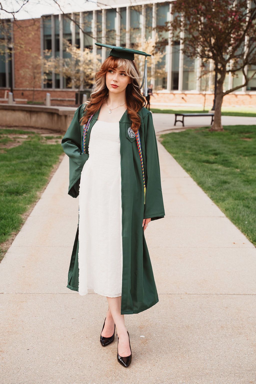 Young woman in green graduation cap and gown over white dress, posing on campus walkway with brick building behind her.
