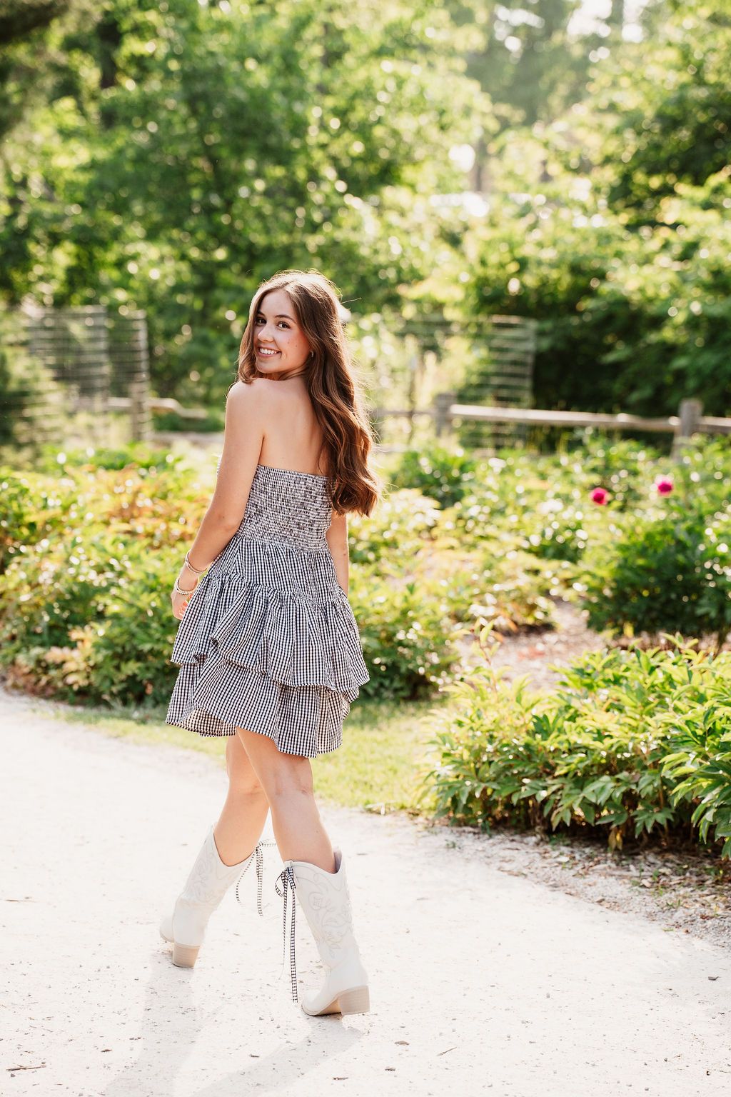 Young woman in striped strapless tiered mini dress and white cowboy boots smiling in a lush garden path.
