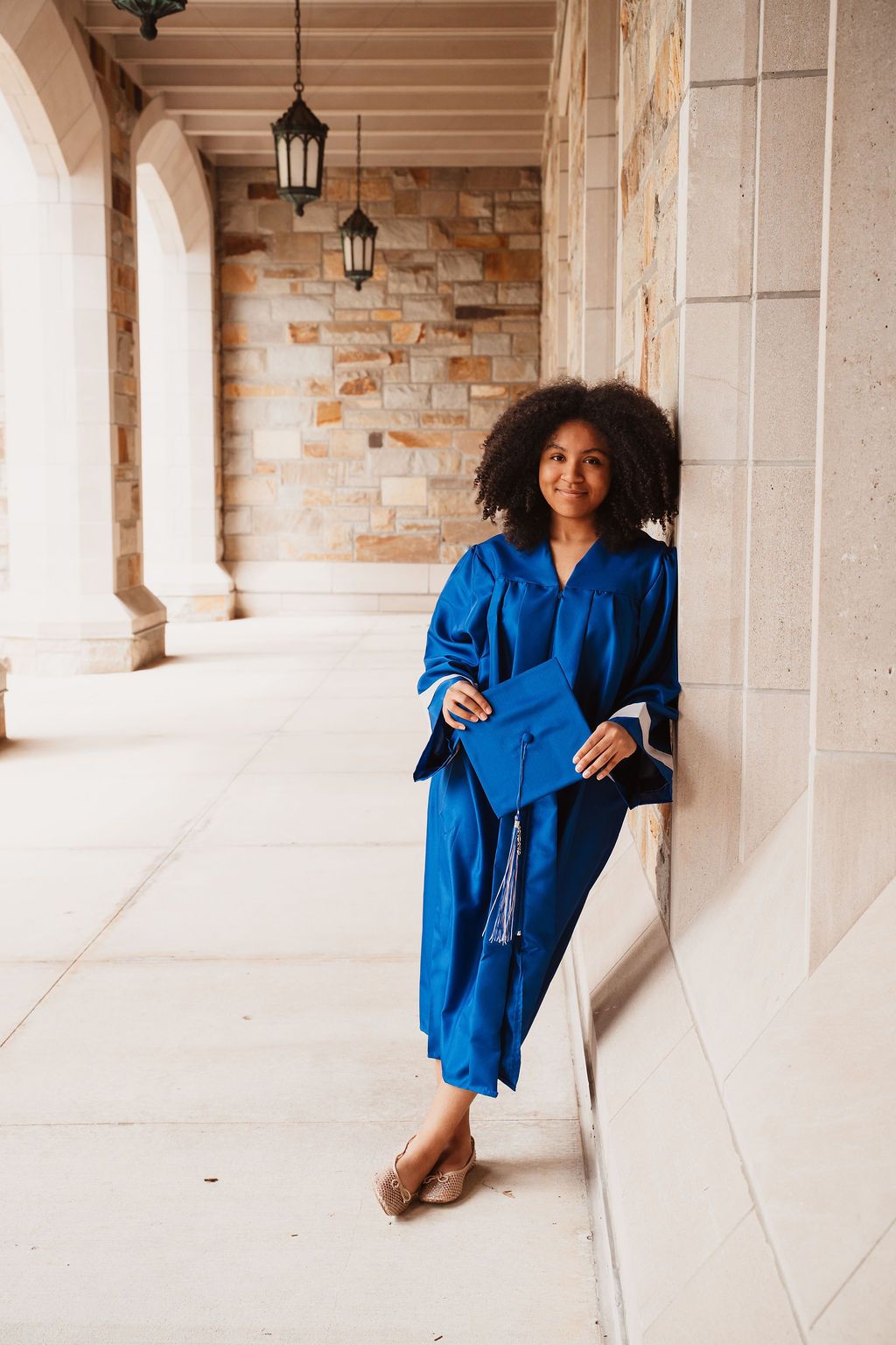 Young woman in blue graduation cap and gown leaning against stone wall in university corridor, smiling confidently.