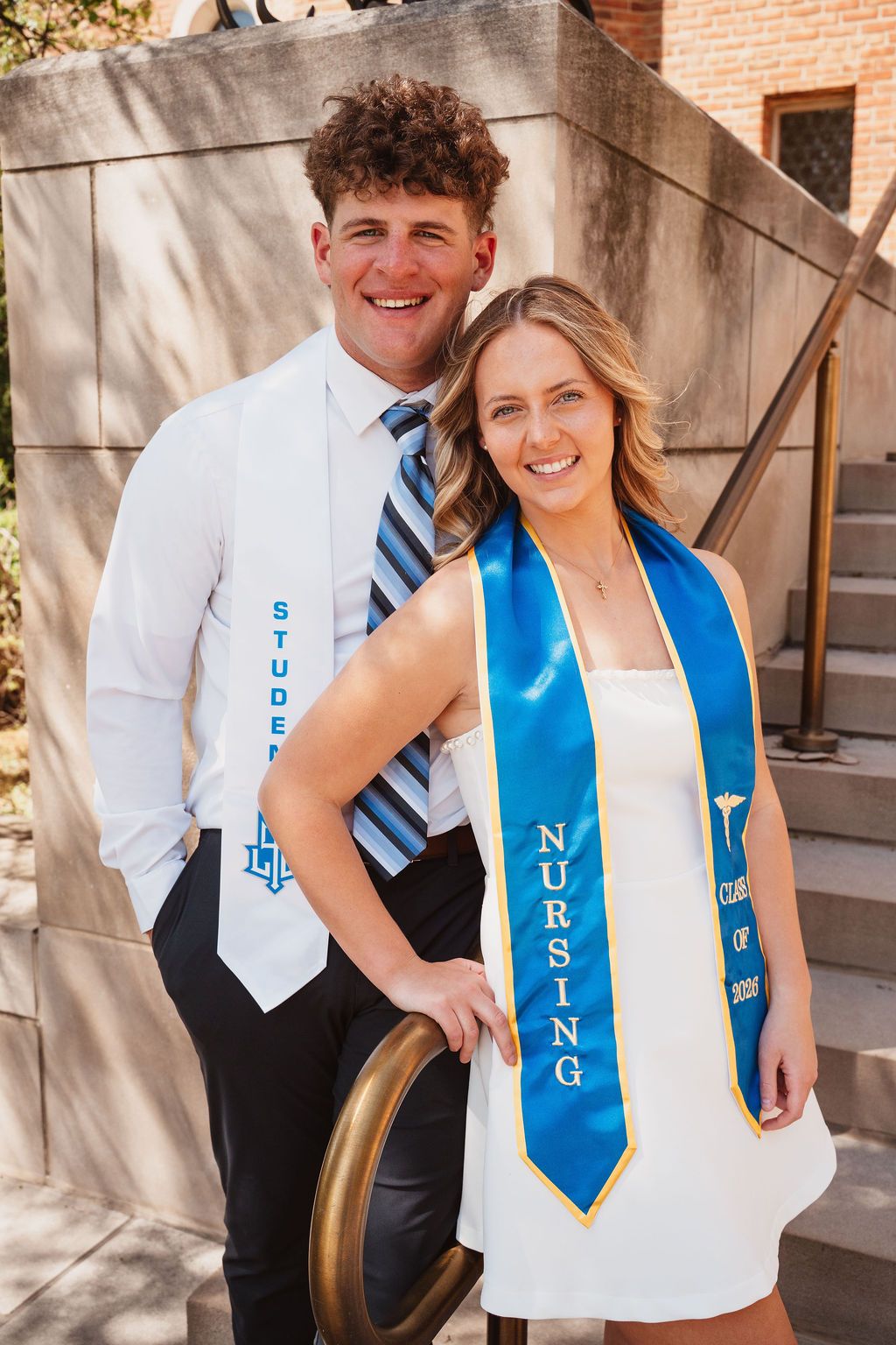 Two smiling graduates pose in white outfits with blue sashes, one reading Nursing, on university steps.