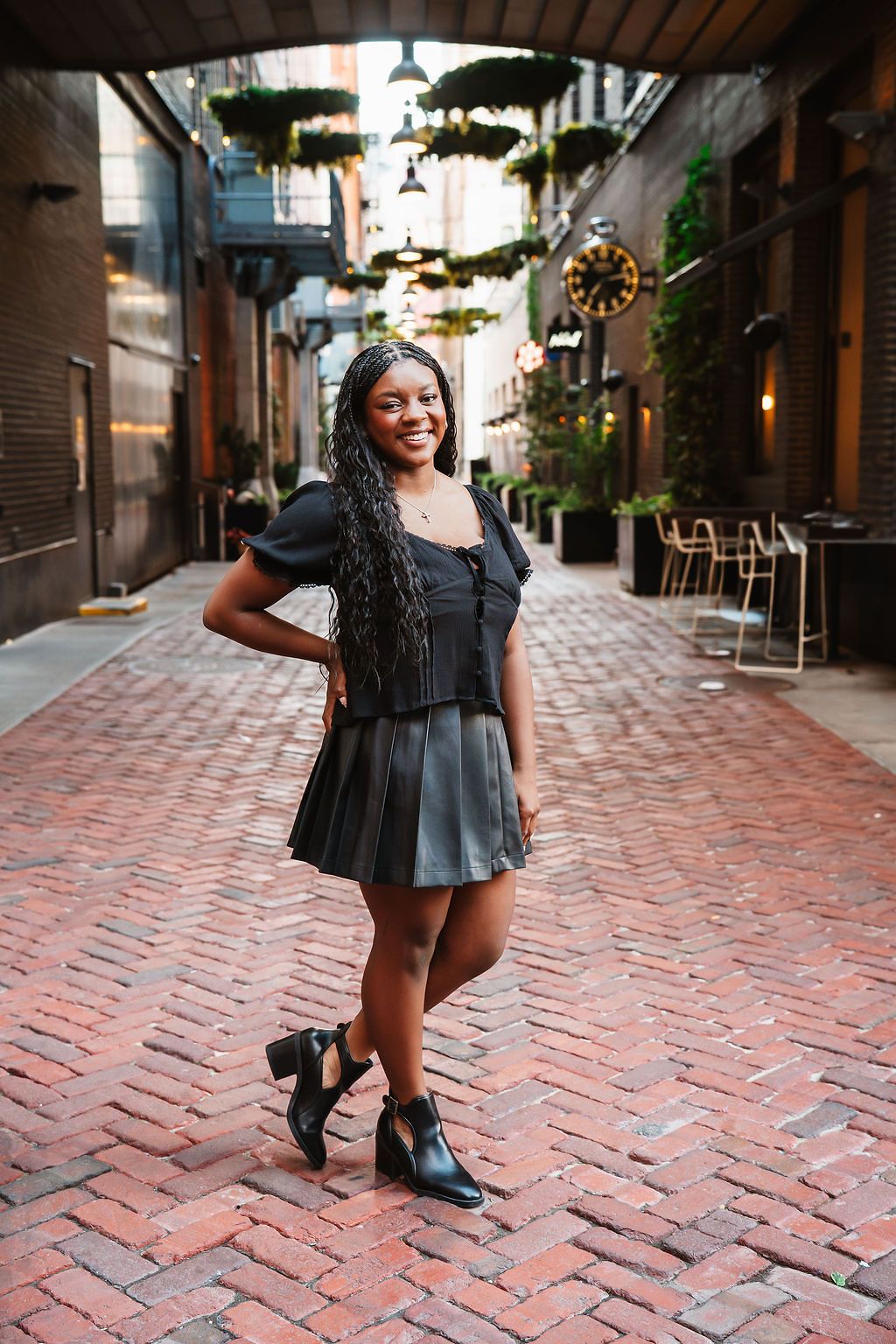 Smiling young Black woman in black pleated skirt and boots posing on brick alley with greenery and string lights.