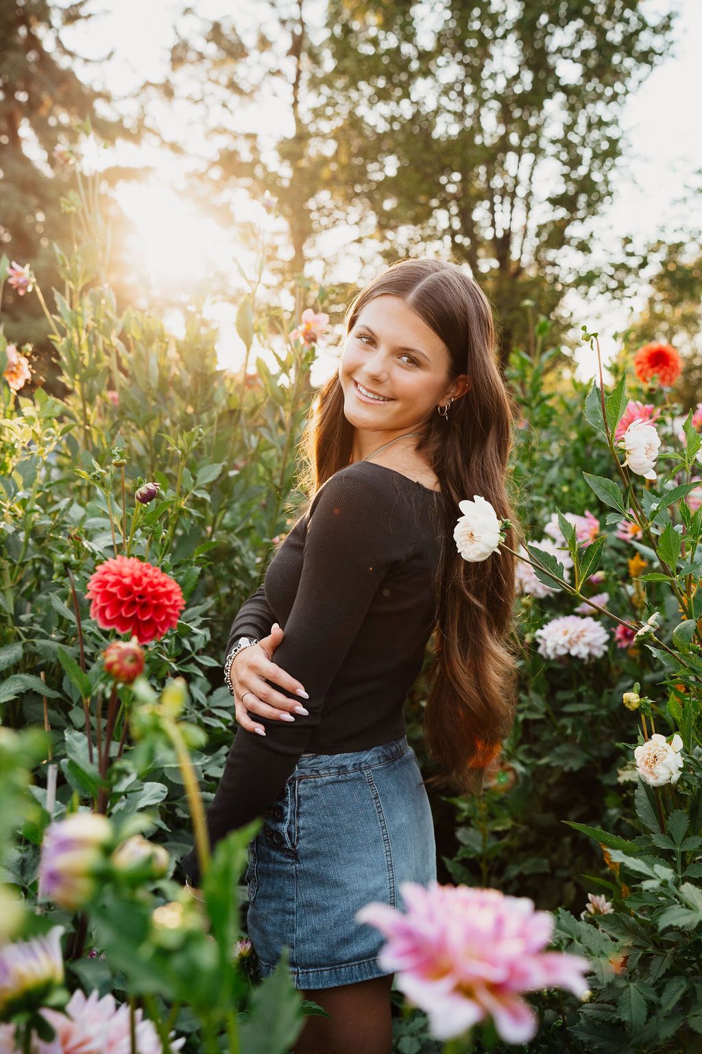 Young woman with long hair smiles in a vibrant flower garden at golden hour, wearing a black top and denim skirt.