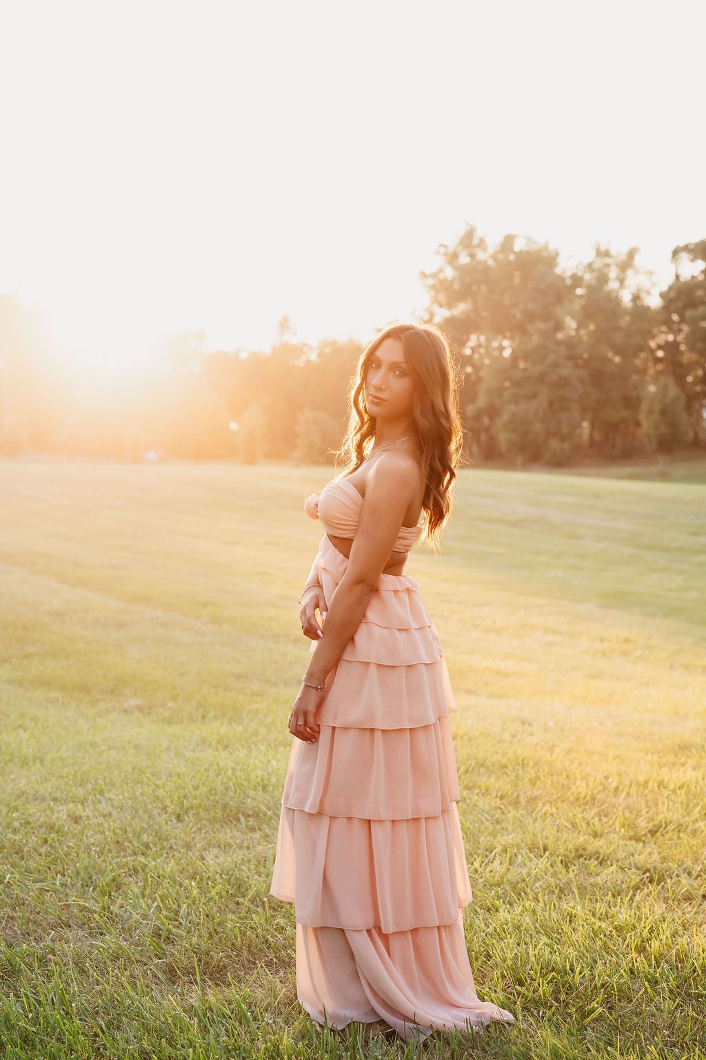 Woman in elegant blush tiered maxi dress standing in golden sunset light on a green field.