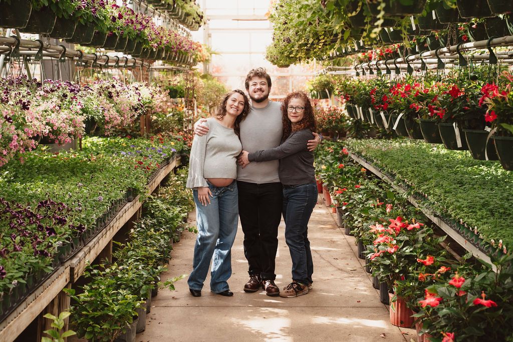 Three people smiling and posing together in a lush greenhouse surrounded by colorful flowering plants.