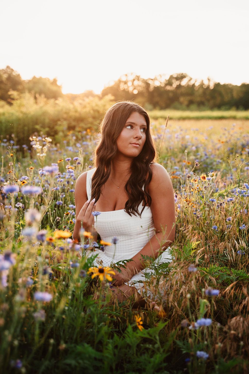 Young woman in white dress sitting in wildflower field at golden sunset hour.
