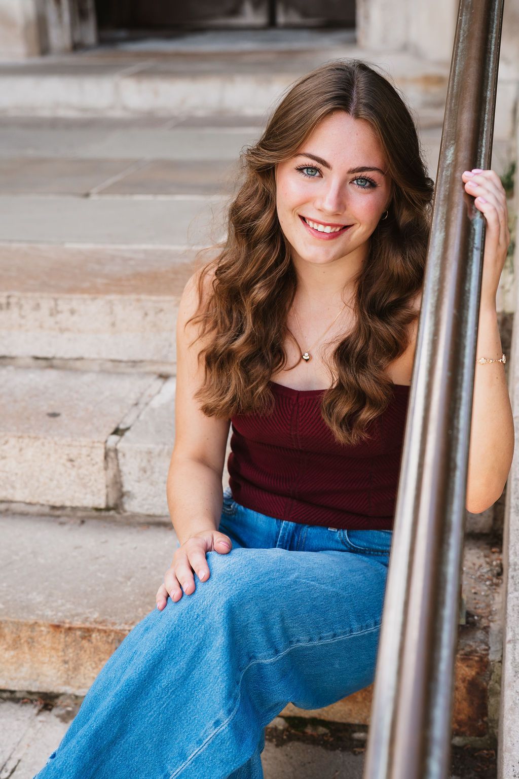 Young woman in burgundy top and blue jeans sits on concrete steps while holding metal railing.