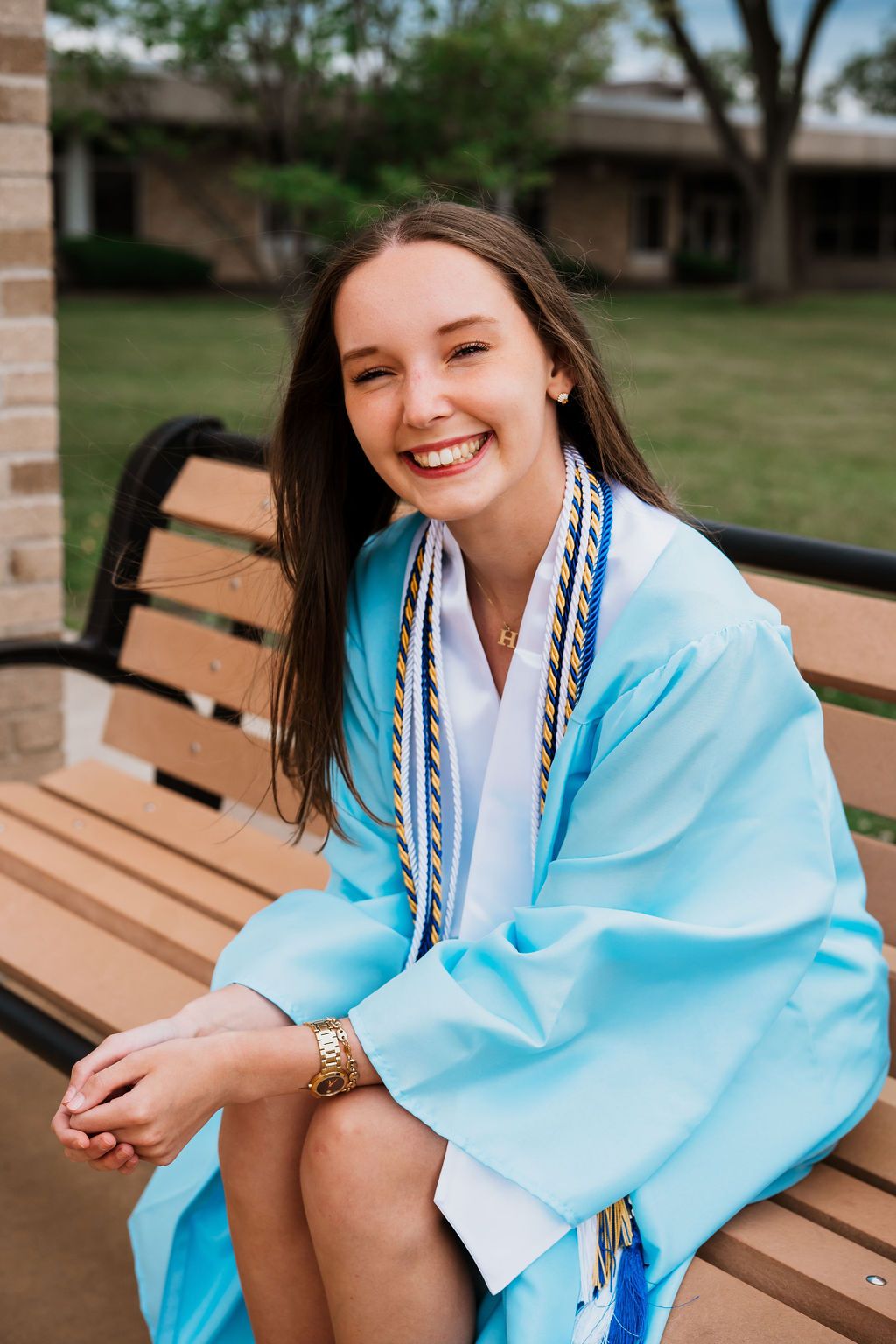 Person wearing light blue graduation gown sits on wooden bench outdoors with bright smile.