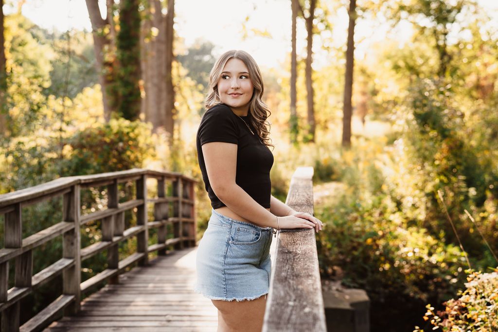 A woman in denim shorts poses on a wooden boardwalk surrounded by golden sunlit trees during sunset.
